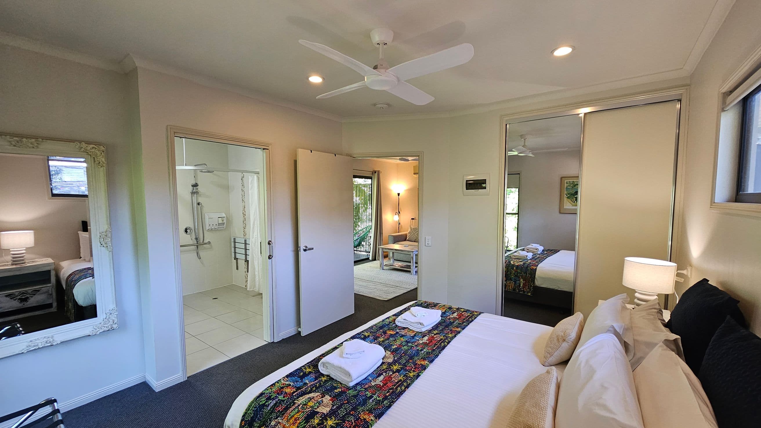 A bedroom with a bed on the right, topped with a colourful runner and neatly folded towels. A bedside table with a lamp is next to it. A large mirror on the left wall reflects the room. The centre shows an open door leading to a bathroom with a shower. Another door opens to a living area with a sofa and a floor lamp. The ceiling has a fan and recessed lights, and the floor is carpeted. A window is visible above the bed.