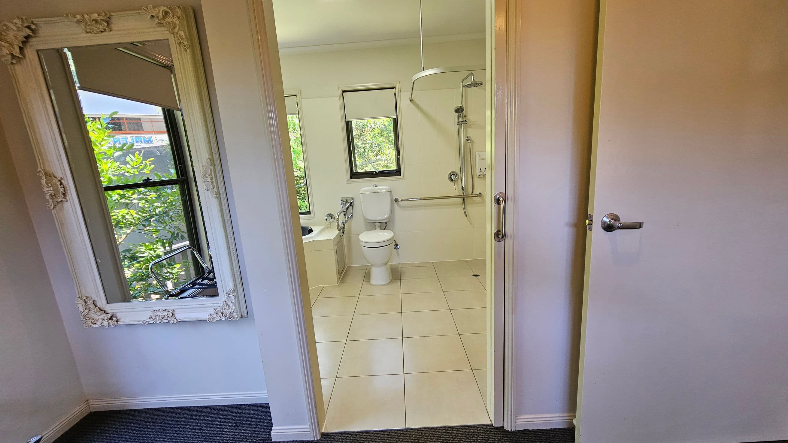 A bathroom with a white tiled floor and a white toilet in the centre. To the left, there's a bathtub with a window above it, partially covered by a blind. On the right, a shower area with a wall-mounted showerhead and a grab bar. The door is open, revealing a room with a large ornate mirror on the left wall, reflecting greenery from outside. The floor in the foreground is carpeted.