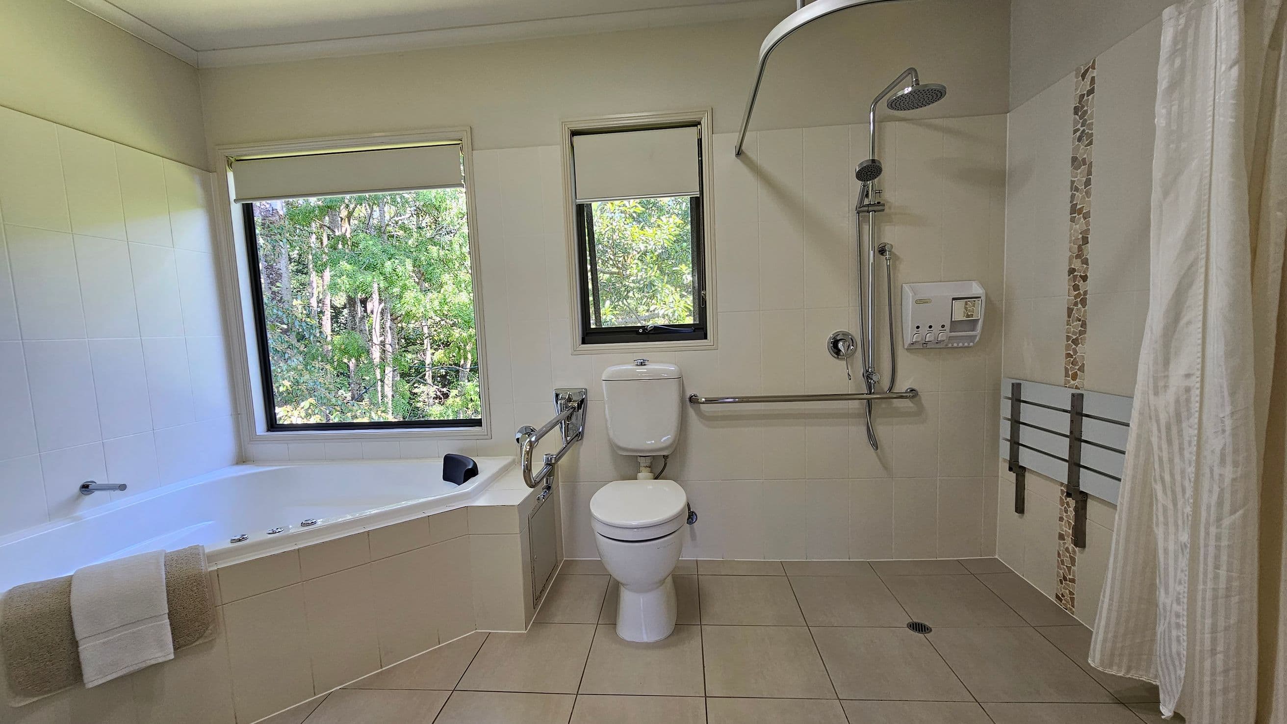 A bathroom with a large corner bathtub on the left, featuring a towel draped over the edge. Two windows with roller blinds provide a view of green trees outside. In the centre, there is a toilet with a support rail. On the right, a shower area with a handheld showerhead, a wall-mounted soap dispenser, and a foldable shower seat. The walls are tiled, and the floor is covered with beige tiles.