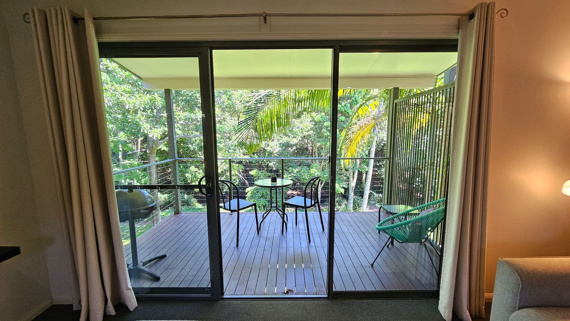 A view through glass sliding doors leading to a wooden deck. On the deck, there's a small round table with two black chairs and a green chair to the right. A barbecue grill is on the left. The background features lush green trees and palm leaves. Inside, beige curtains frame the doors, and a grey sofa is partially visible on the right. A lit wall lamp is above the sofa.