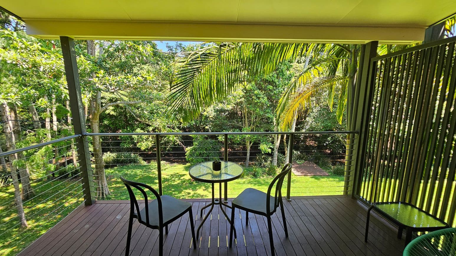 A wooden deck with a small round glass table and two chairs. A potted plant sits on the table. The deck is surrounded by a metal railing with cable wires. On the right, there's a vertical slatted screen. In the background, lush green trees and a well-maintained lawn are visible. A large palm frond hangs over the railing. The ceiling is light-coloured, and the floor is made of dark wooden planks.