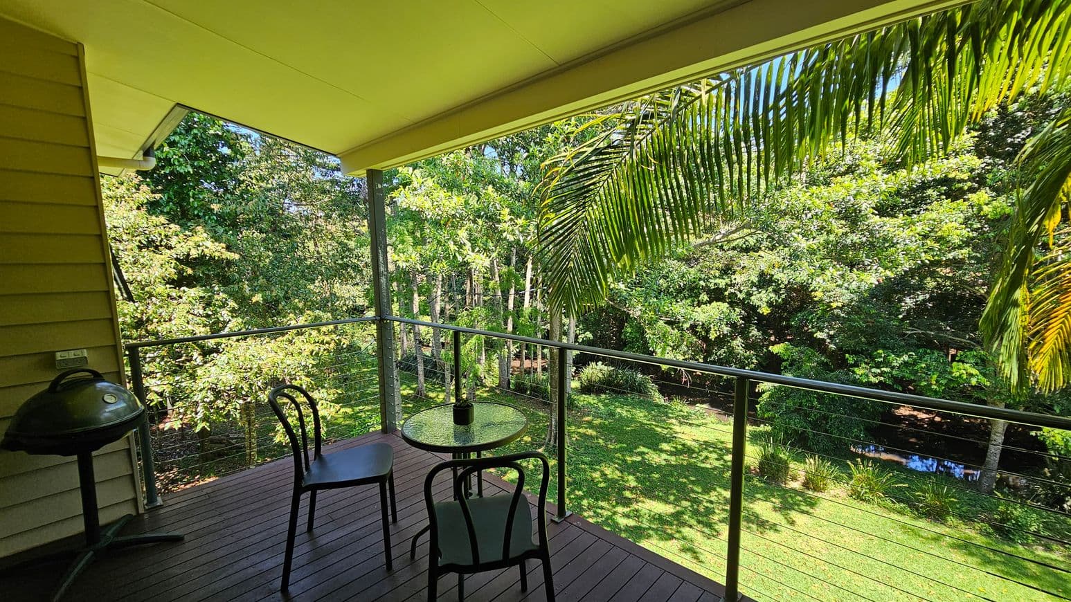 A wooden deck with a small round glass table and two black chairs. On the left, there's a barbecue grill next to a yellow wooden wall. The deck is bordered by a metal railing with cable wires. In the background, lush green trees and a well-maintained lawn are visible, with a small stream running through the garden. The ceiling is a light yellow, matching the house exterior. Palm fronds hang down from the top right corner.