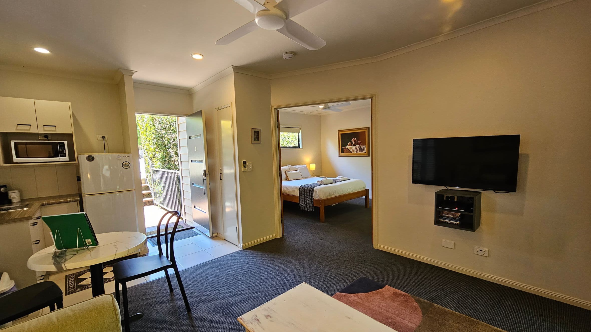 A compact living space with a kitchenette on the left, featuring a microwave, fridge, and cabinets. A small round table with chairs is nearby. The entrance door is open, revealing greenery outside. In the centre, a doorway leads to a bedroom with a double bed, bedside lamp, and a painting on the wall. On the right, a wall-mounted TV with a media player below. The floor is carpeted, and a ceiling fan is visible.