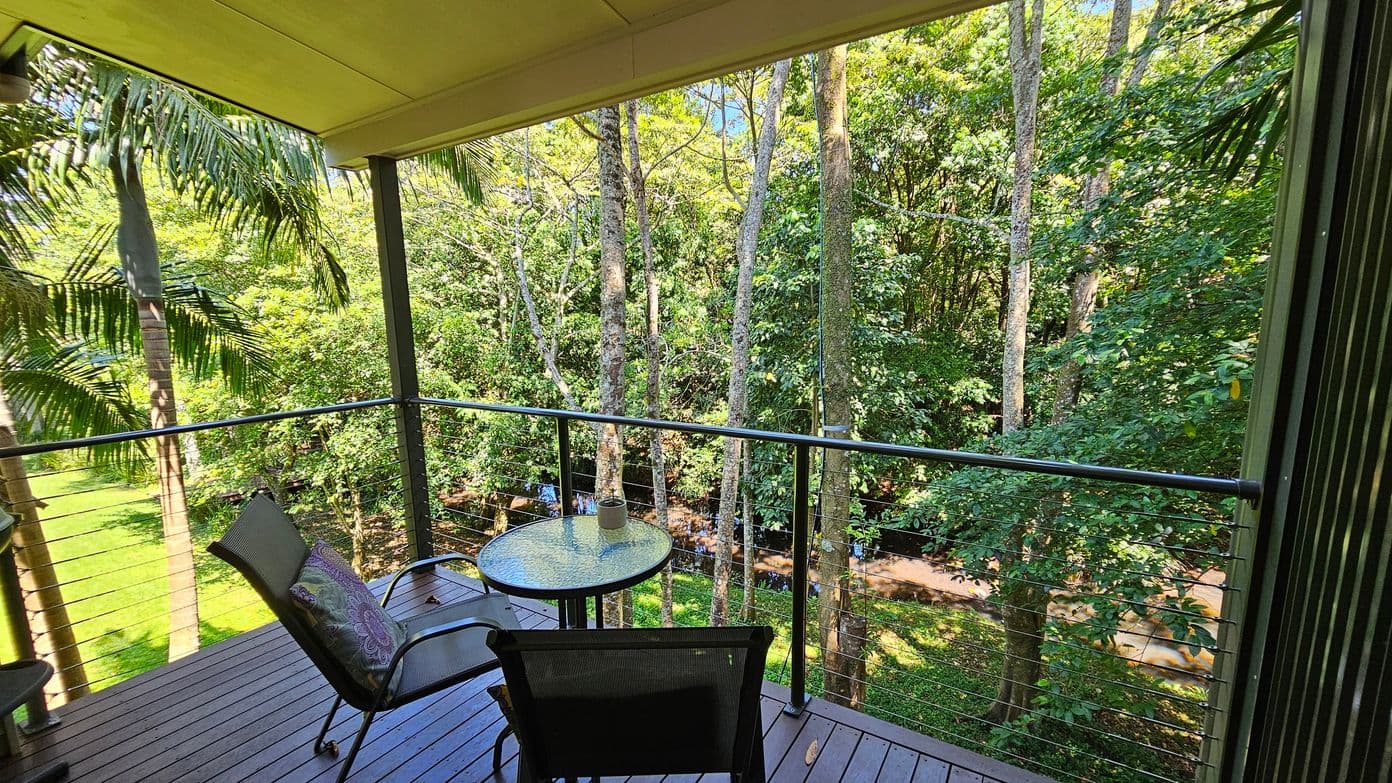 A wooden deck with two cushioned chairs and a round glass table, set against a backdrop of lush green trees. The deck is enclosed by a metal railing with wire cables. Tall palm trees are visible on the left, and a small stream runs through the forested area in the background. The ceiling of the deck is light-coloured, providing shade to the seating area.