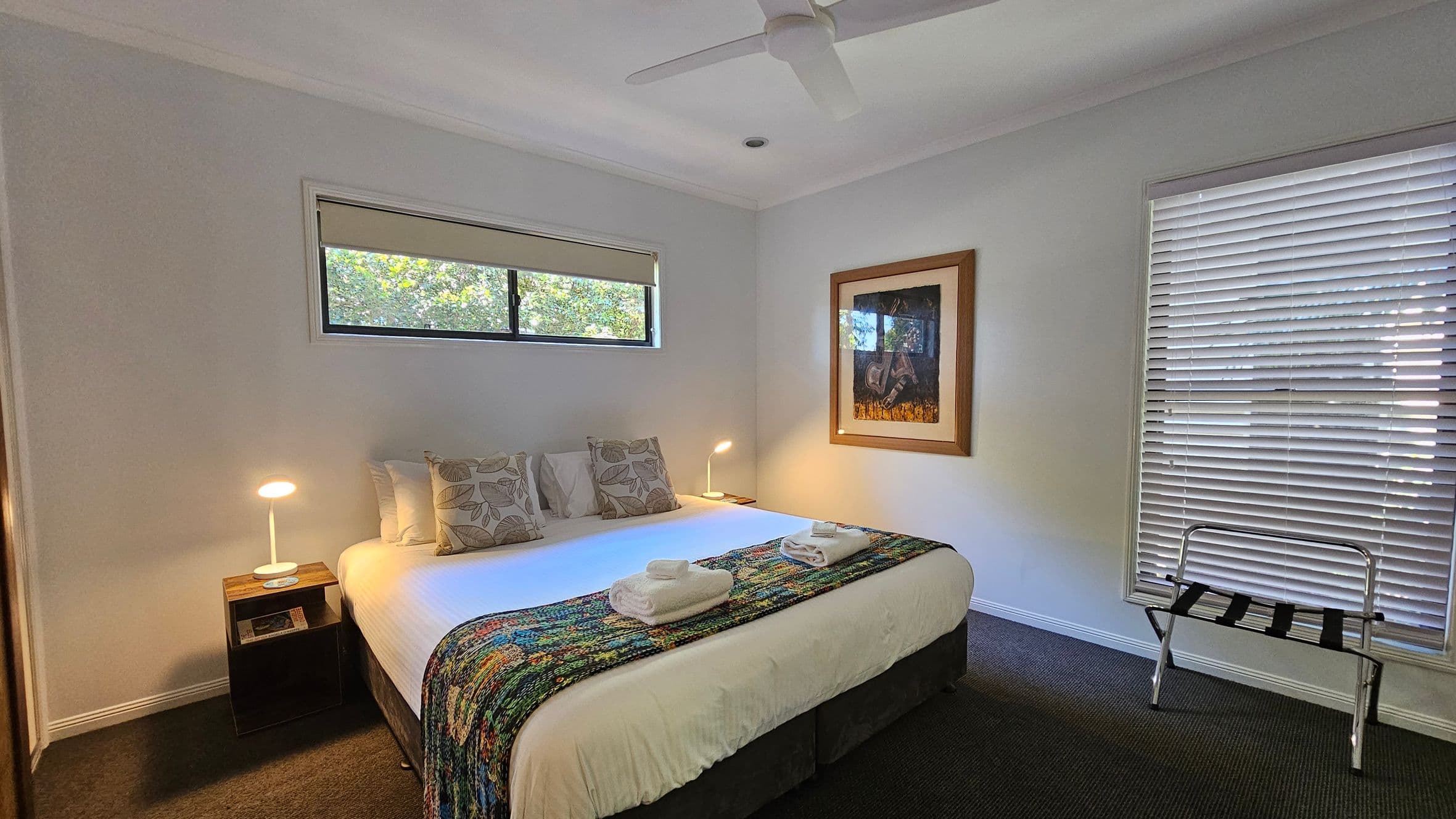 A bedroom with a large bed in the centre, adorned with a colourful throw and two folded towels. Two bedside tables with lamps flank the bed. Above the bed is a narrow window with a blind. On the right wall, a framed picture hangs above a luggage rack. A window with closed blinds is also on the right. The room has a ceiling fan and a dark carpeted floor.