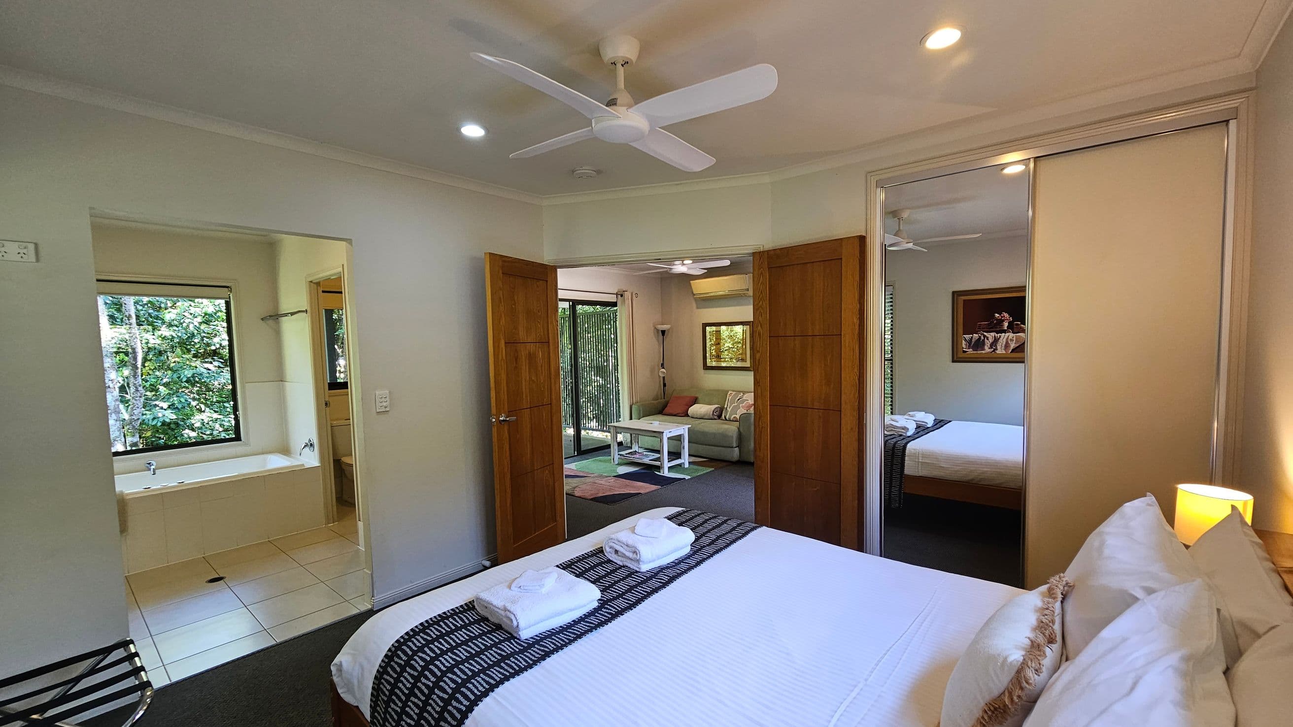 A bedroom with a neatly made bed on the right, topped with two folded towels. A mirrored wardrobe reflects part of the room. In the centre, wooden double doors open to a living area with a sofa, coffee table, and a floor lamp. On the left, an open doorway leads to a bathroom with a bathtub and a window showing greenery outside. The ceiling has a white fan and recessed lights.