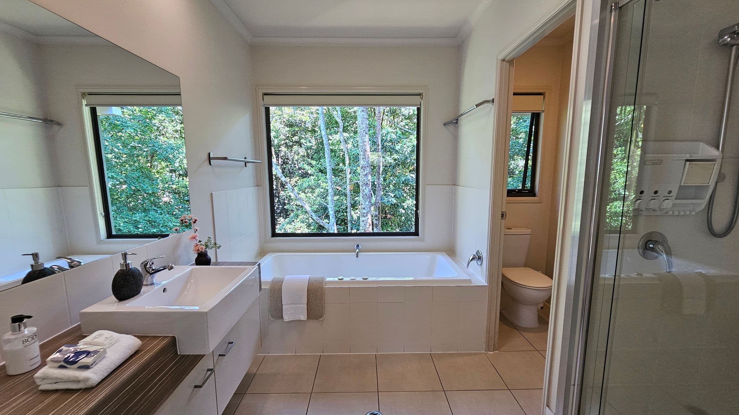 A bathroom with a large window in the centre, offering a view of lush green trees. Below the window is a white bathtub with a towel draped over the edge. To the left, a white sink with a wooden countertop holds soap dispensers and a folded towel. A large mirror is above the sink. On the right, a glass shower enclosure is visible. The floor is tiled in a light beige colour, and a small room with a toilet is seen through an open door