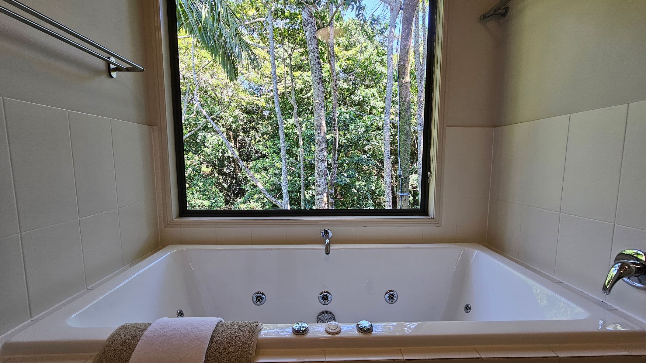 A white bathtub with a chrome faucet is positioned beneath a large window. The window offers a view of lush green trees with slender trunks and dense foliage. The walls surrounding the bathtub are tiled in white. A towel is neatly placed on the edge of the tub. A metal towel rack is mounted on the left wall.