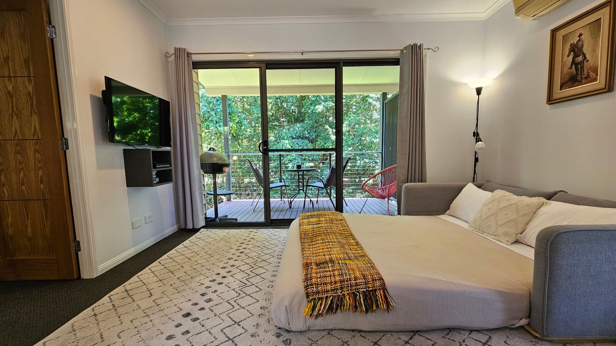 A cosy room with a bed on the right, covered with a beige blanket and a colourful throw. Above the bed is a floor lamp and a painting of a horse and rider. On the left, a wall-mounted TV is above a small shelf. The floor has a patterned rug. A sliding glass door in the centre leads to a balcony with a small table, chairs, and a barbecue grill. The balcony overlooks lush green trees. The ceiling is white with a mounted air conditioning unit.