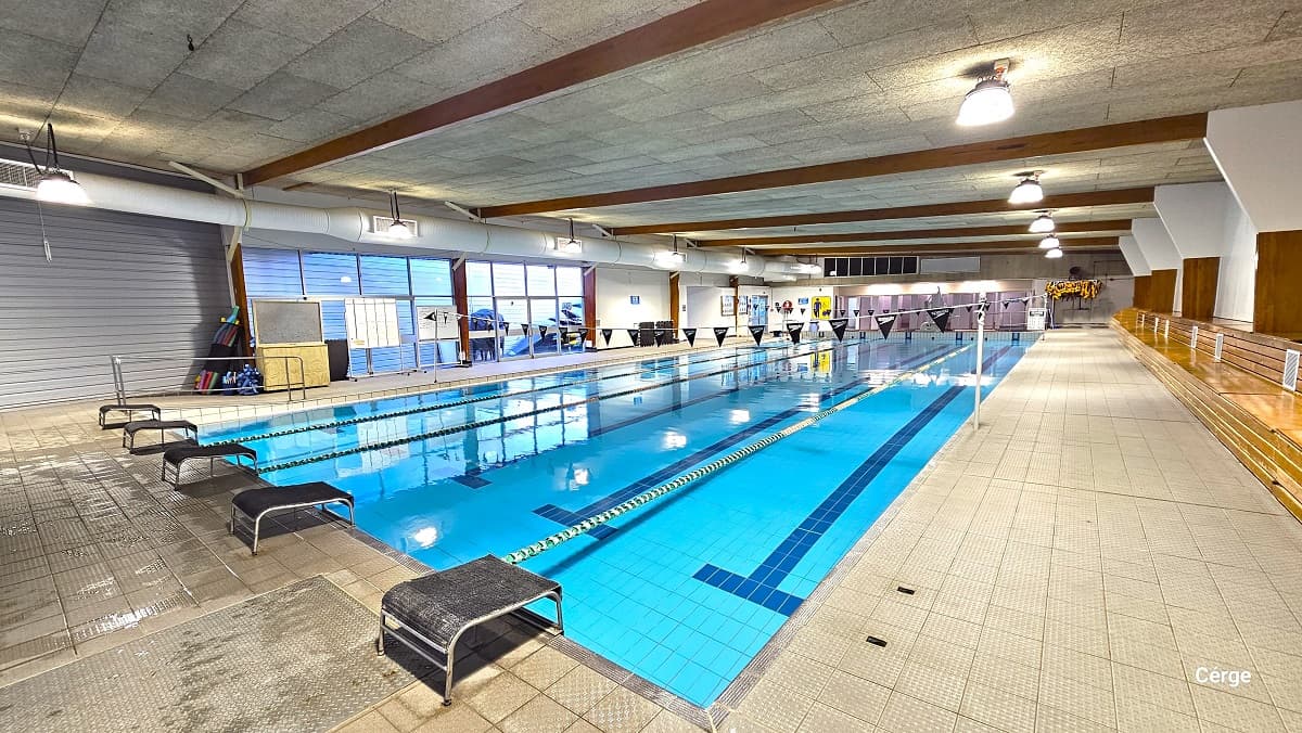 Pool deck near the 25m lap pool leads to the manually operated glass doors with a green exit sign on top. On the left is the blue-tiered seating along the wall, and on the right is a roll of blue waterproof tarpaulin wrapped around a metal tube. There is a yellow floating device on the white behind the tarpaulin and a NO BOMBING and Masterton Swimming Club signboard.