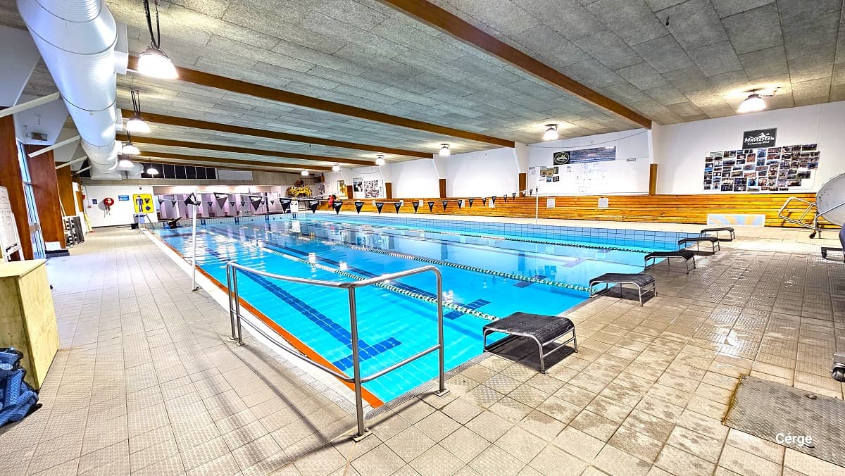 Closer view of the 25m program pool on the handrail side. The program pool can be distinguished by the grey tile ceiling with brown wooden roof beams. The area is well-lit by pendant lights lined on the sides of the ceiling. On the white wall are several photo cards just below the Masterton swimming club logo.