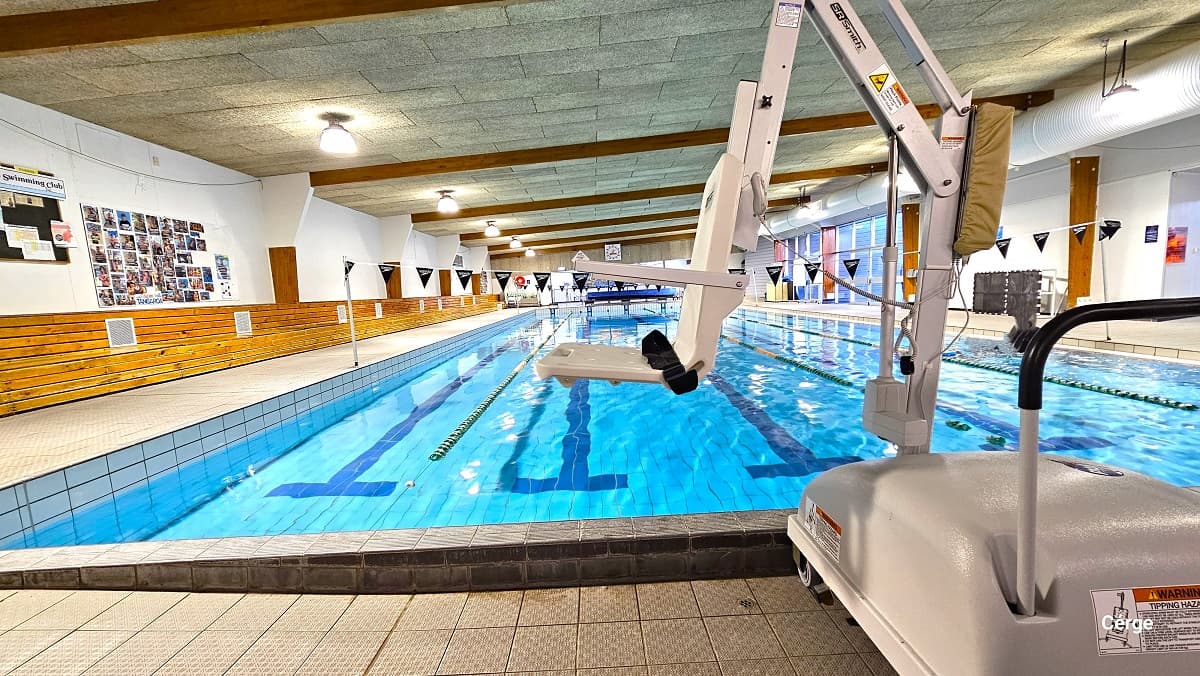 White pool hoist in the 25m Program Pool at Trust House Recreation Centre. On the left wall, there is a black bulletin board with papers, and next to it are several photo cards arranged in a square shape.