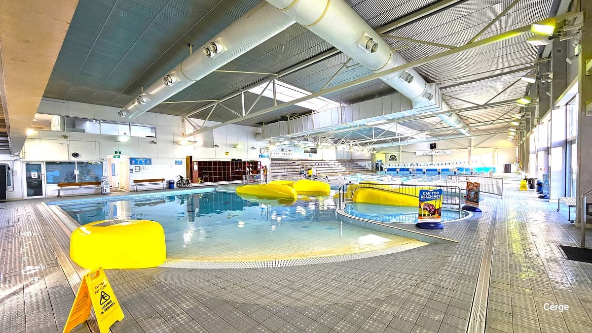 Toddler pool has shallow water with an irregular yellow shape placed on the pool edges. Next to it is the spa and sauna pool, with a yellow waterslide occupying most of the space at the center, while further beyond is the 25m lap swimming pool. The aquatic centre has grey tiles with circular bumps for foot friction to avoid slipping, a corrugated roof pattern, and a large, white oval duct for ventilation. On the left is a brown wooden shelf for bags.