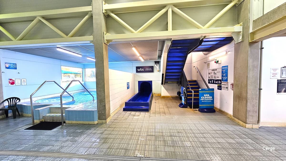 The Sauna pool and blue speed slide are separated by a white-tiled wall at Trust House Recreation Centre. The saun pool has a black mat placed before the two-step stair with steel handrails and a black plastic chair with an armrest. Further to the back wall is a glass window. On the right is the end of the 8-lane speed slide and a blue stair leading to the spa. At the bottom of the stairs is a “spa closed for cleaning” blue signboard.