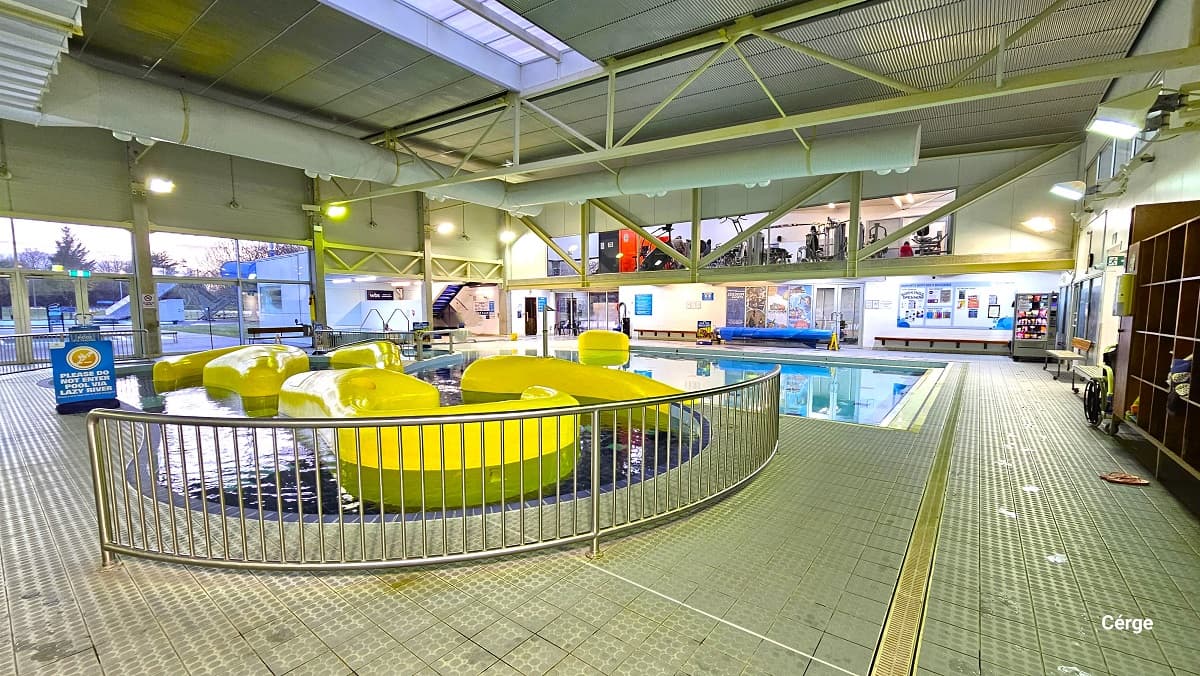 View of the leisure pool with yellow irregular shapes in the centre. There is a short steel fence on both sides, while on the back of the pool is a blue sign board that says, “Please do not enter the pool via lazy river.” Opposite this position is the exit glass doors to access the aquatic amenities outdoors. The upper gymnasium can be seen through the glass windows and two wooden wall-mounted seating beside the vending machine.