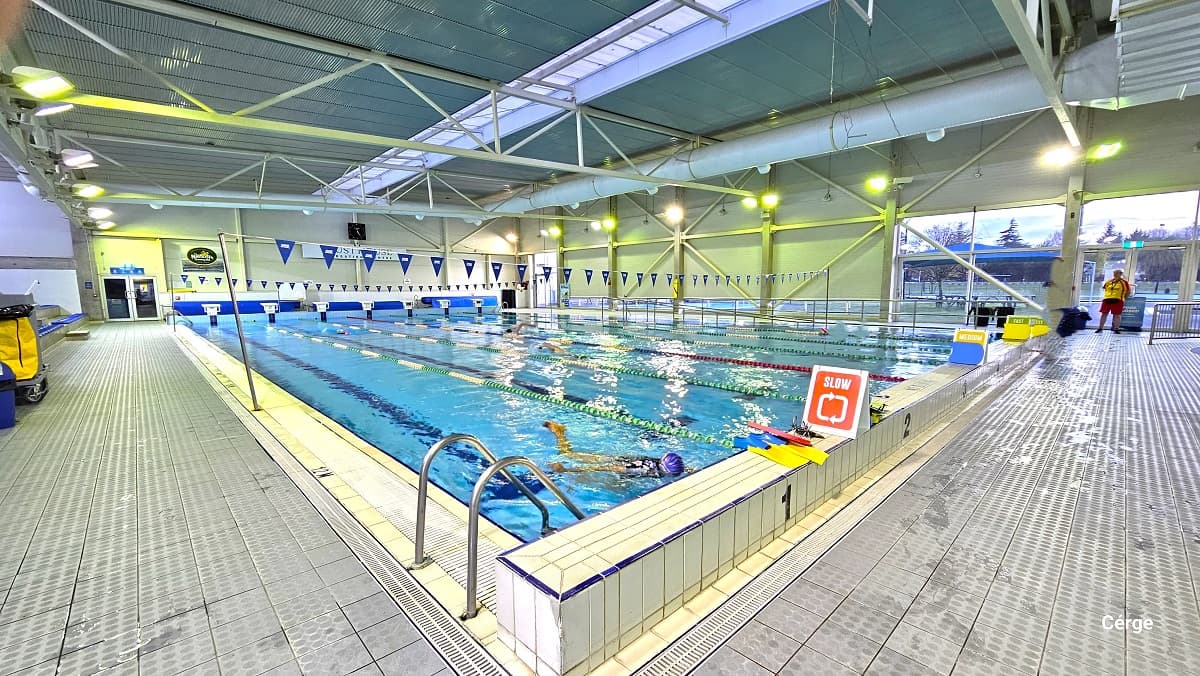 25m lap swimming pool inside the Aquatic Centre at Trust House Recreation Centre. The lanes are divided by green, white, red, blue, and yellow floating rope lanes. The pool is well-illuminated by several wall lighting fixtures attached to the grey roof beams. The left side has pool ladders marked by inverted U-shape metal frames, while to the right is ramp access with steel handrails. The pool has slow, medium, and fast lane signboards at the end and white diving blocks at the start.