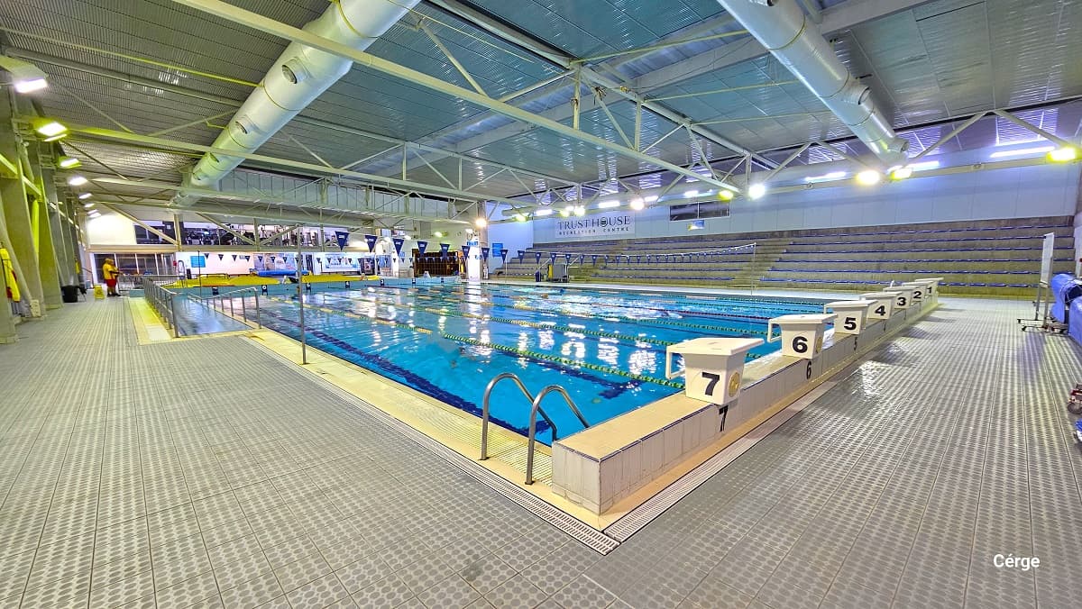 A closer look at the numbered 1-7 white diving blocks and ramp access of the 25m lap swimming pool. There are two strings of blue triangular flags wrapped around two grey metal poles. The name of the center is detailed on the large white signboard on the wall in the multi-tiered seating area with blue seats opposite this position. Found close to the diving blocks is a mobile whiteboard. Lifeguards can be seen roving the area wearing the yellow and orange uniforms.