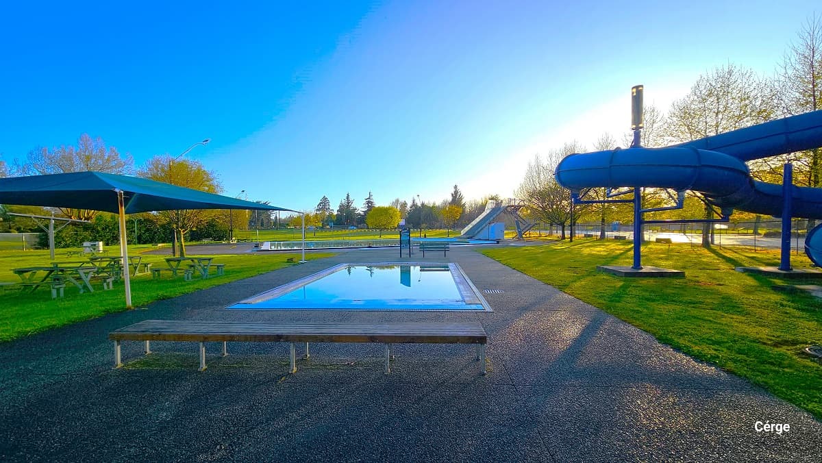 Outdoor 2m dive-well at Trust House Recreation Centre with a natural stone tile. On the left side is a seating area with picnic tables and a blue sunshade. On the right side is the 8-lane speed tubular slide, while to the back is the 25m lido pool. The landscape surrounding the amenities is filled with well-maintained grass and a vivid shade of green. The outdoor also offers the natural environment of tall trees, rays of sun, and calm blue sky, perfect for relaxation.