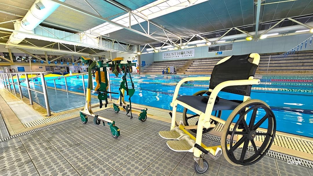 Accessible aquatic wheelchair and mobile hand support with green and orange straps at the entrance of indoor 25m lap pool. The aquatic wheelchair has a blue cushion seat and a black support with a light brown footrest.