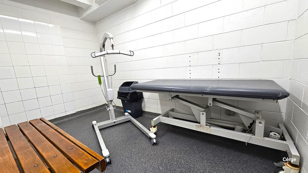 Accessible changing room at Trust House Recreation Centre. It has a granite-like tile floor style and a white-tiled wall. Found inside the room is a wooden wall-mounted seat, a changing table with dark blue cushion, and a white patient lift. On the left side of the changing table is a grey garbage bin with a black garbage bag.