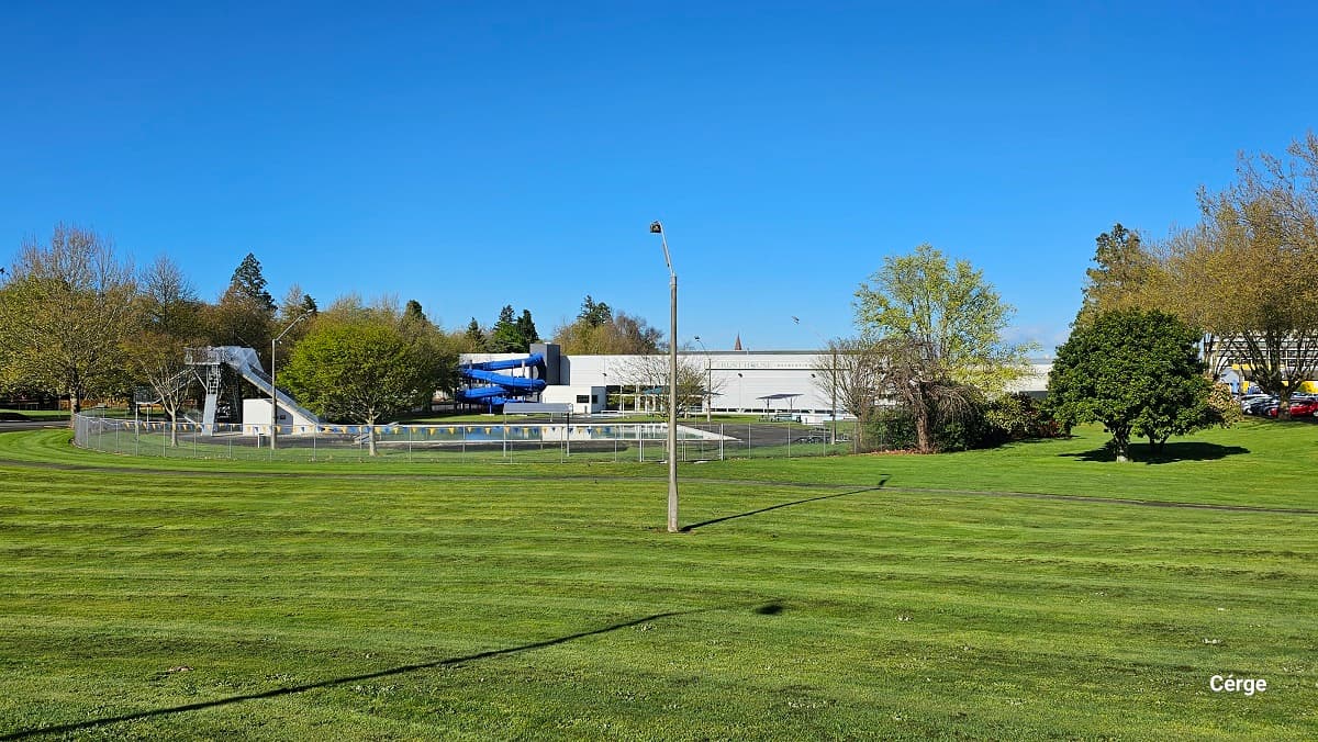 A large open green field with well-maintained grasses surrounds the Trust House Recreation Centre. In the middle of the field is a lamp post. Protruding right outside the facility is a spiraling blue water slide, and a tower with metal stairs, an outdoor pool with a blue canopy shade which are all enclosed by a chain-linked fence with blue and yellow triangular flags circling around the property.