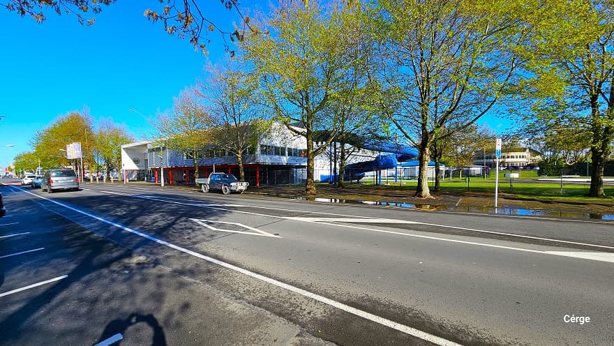 View from the right side angle of Trust House Recreation Centre from Dixon Road. Roadside parking along Dixon can be distinguished by the white painted lines. The road also has white diamond markings with slight horizontal lines. The spiraling tube of the blue water slide is situated on the right side. Tall trees with great abundant foliage offer shade to the cars parked under them. The road is quite busy, with cars passing frequently as the center is set within the commercial area.