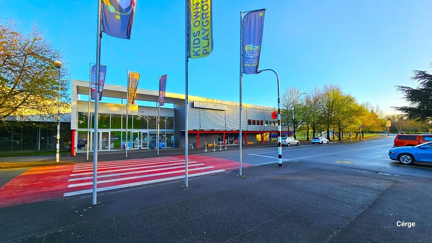 Step-free access marked by flag poles from the car park with accessible parking spaces on the right marked by a yellow sign for persons with mobility disabilities. From the step-free access is a red pedestrian crossing leading to the automatic glass sliding entrance doors. Cars are parked in the further right corner in front of the facility. Tall trees with large canopy cover can be seen in the surrounding area of the facility.