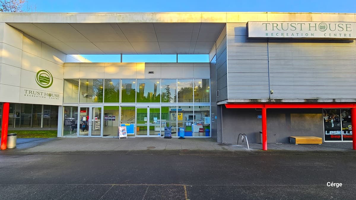 Front view of Trust House Recreation Centre. The facility is a large two-story building with white-framed glass windows, corrugated iron façade, and red, green, and silver features. The name of the facility is detailed on the left wall with the brand logo on top of the name and to the right corrugated wall in a large rectangular sign board. There is a single bike rack on the right side just before the entrance. A number of advertising posters are placed on the glass wall of the main entrance.