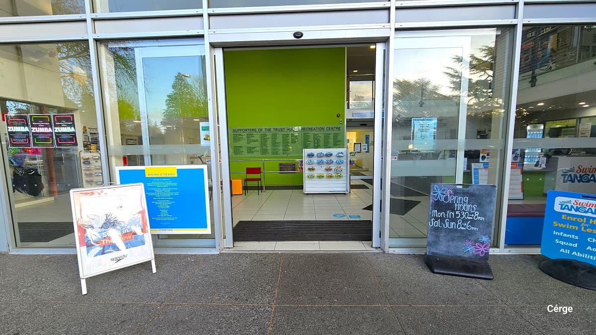 Entry to the Trust House Recreation Centre is through the automated sliding glass doors with white metal frames. In front of the entrance door, on the right side, is a chalkboard indicating work hours, and beside it is a blue sign board for swimming lessons. Other advertisement signboards can also be seen on the left of the front glass wall. Small blocks of white painted wood with swimwear and accessories can be seen right after the entrance doors.