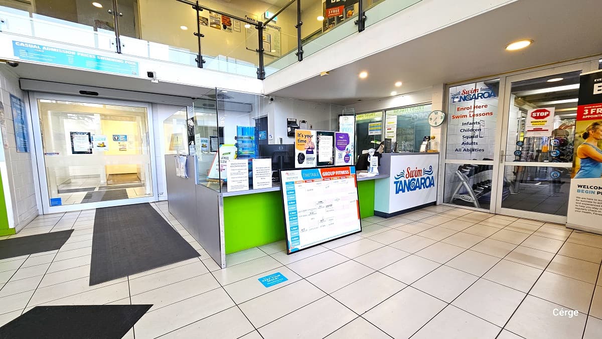 Front side angle of the reception desk is light green. In front of it is a large white sign board for the timetables and group fitness information. The Swim Tangaroa logo in red and blue is painted on the other white table. Next to it, on the upper white wall, is a white clock. The entry to the lift and stairs and heavyweights area is through a large glass sliding door to left and right of the reception desk, respectively. Advertisements of different sizes are scattered in the reception area.