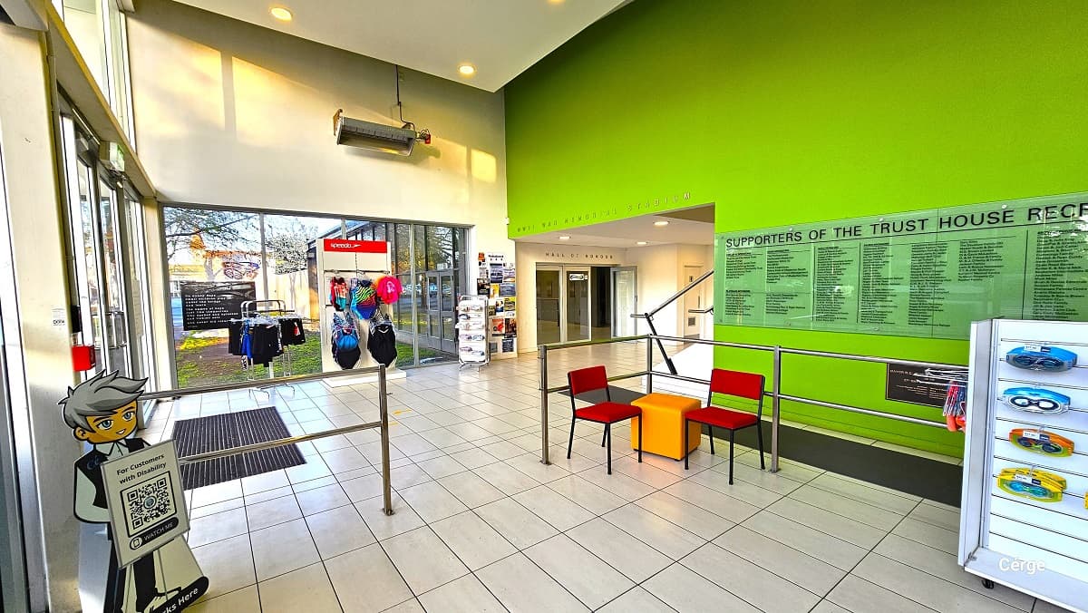 View of the small retail display after passing through the automated glass sliding doors. Separating the entryway and the reception area is a steel handrail enclosing the Cérge standee holding the QR for customers and two red metal chairs with an orange sofa cube stool. Next to the glass wall to the left, from the entrance door, is the swimwear display, while straight ahead is the Hall of Honor entrance glass doors.