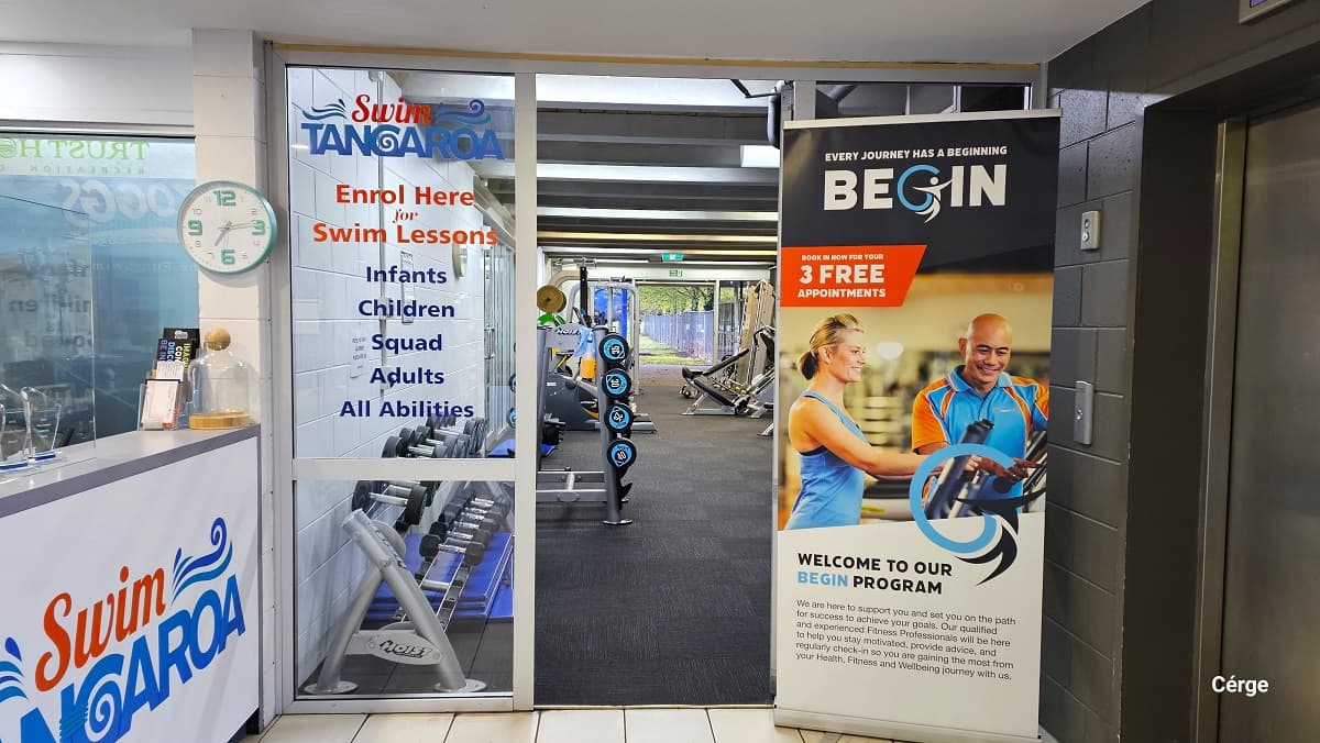Ground level gymnasium entrance at Trust House Recreation Centre. Entry is through manually operated glass entrance doors with white metal frames right next to the reception desk. On the right side is a large advertisement for their Begin Program. The left glass door has a Swim Tangaroa logo with ‘enroll here for swim lessons’. Its gymnasium has grey carpet tiles. Near the entrance doors are black dumbbells.