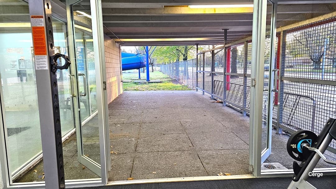 Manually operated glass exit doors of the ground-level gymnasium at Trust House Recreation Centre. The pathway leading outside is made of large natural stone tiles. On the right side is a wire fence, while straight ahead is the large blue spiraling waterslide.
