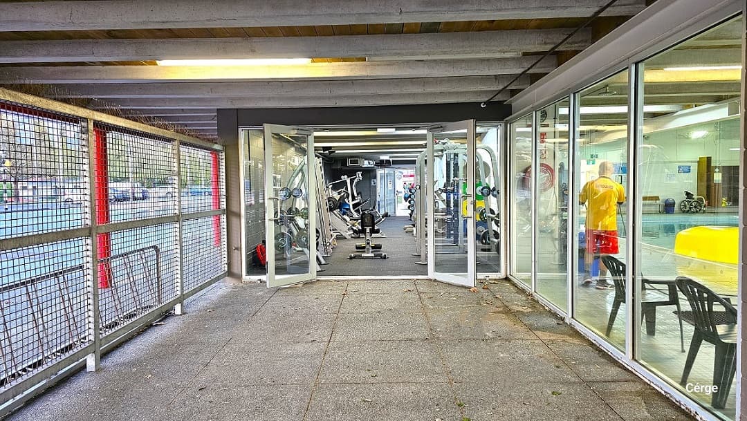 View when standing from the natural stone tiles a few meters away from the manually operated exit glass doors. On the right side is a glass window that has a view of the aquatic center pool. On the left side is the wire fence with a view of the car park spaces outside the centre.