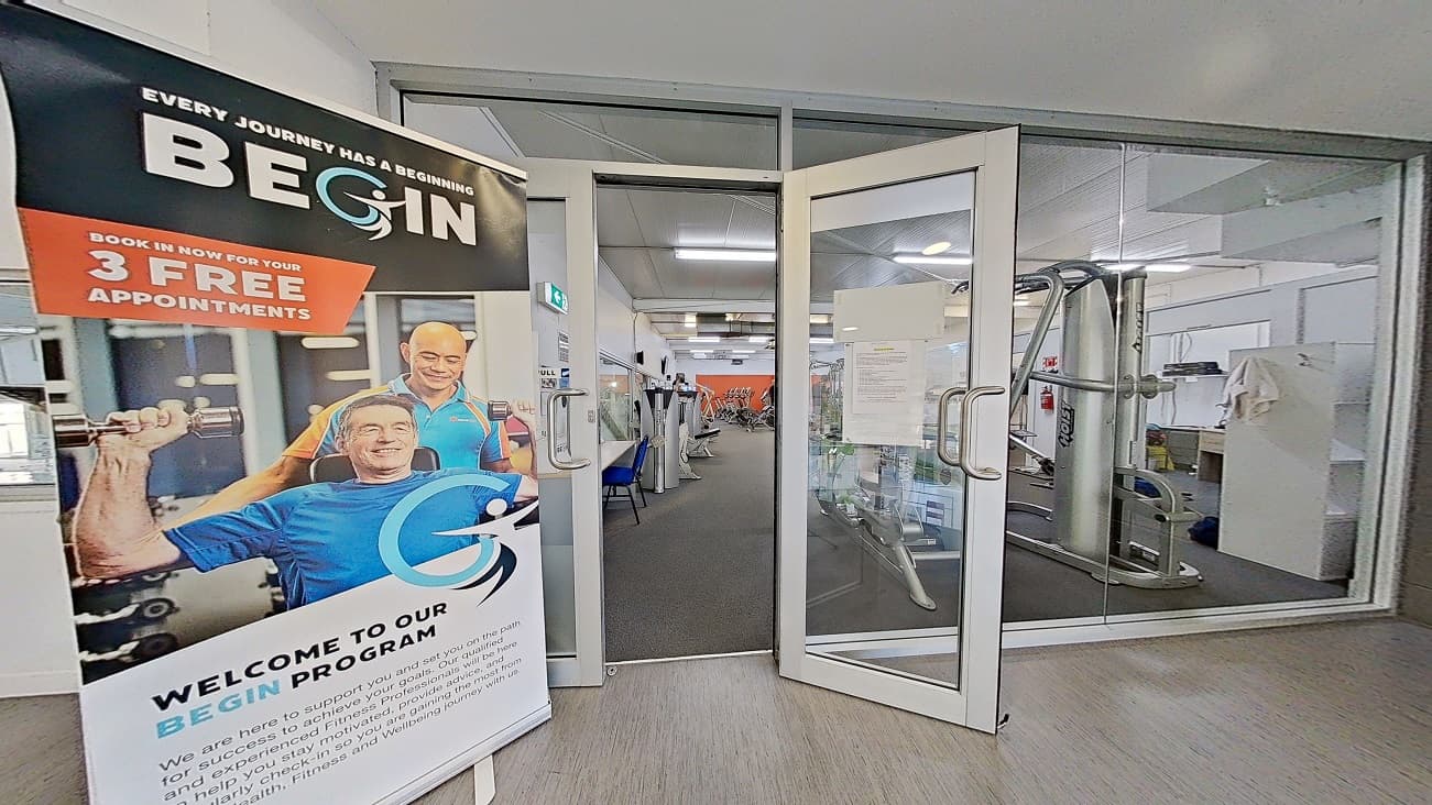 Upper-level gymnasium at Trust House Recreation Centre. The upper level has smooth grey tiles with a yellow foot icon on the middle of the floor leading to the manually operated outward-opening glass doors and glass with steel railing. ‘Begin Program signboard’ is placed on the left side of the entrance glass doors. Behind it is a black chalkboard with “I don’t sweat, I sparkle” writing.