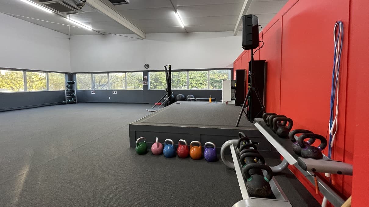 Studio 1 at the upper level of Trust House Recreation Centre. The studio has a large open space with grey carpet tiles. There is a small grey stage in front, along with speakers on top of it. On the right side of the stage are kettle bells of different colors, weights, and sizes. The front wall is painted in red while the rest is half painted in grey and white. It has small glass windows.