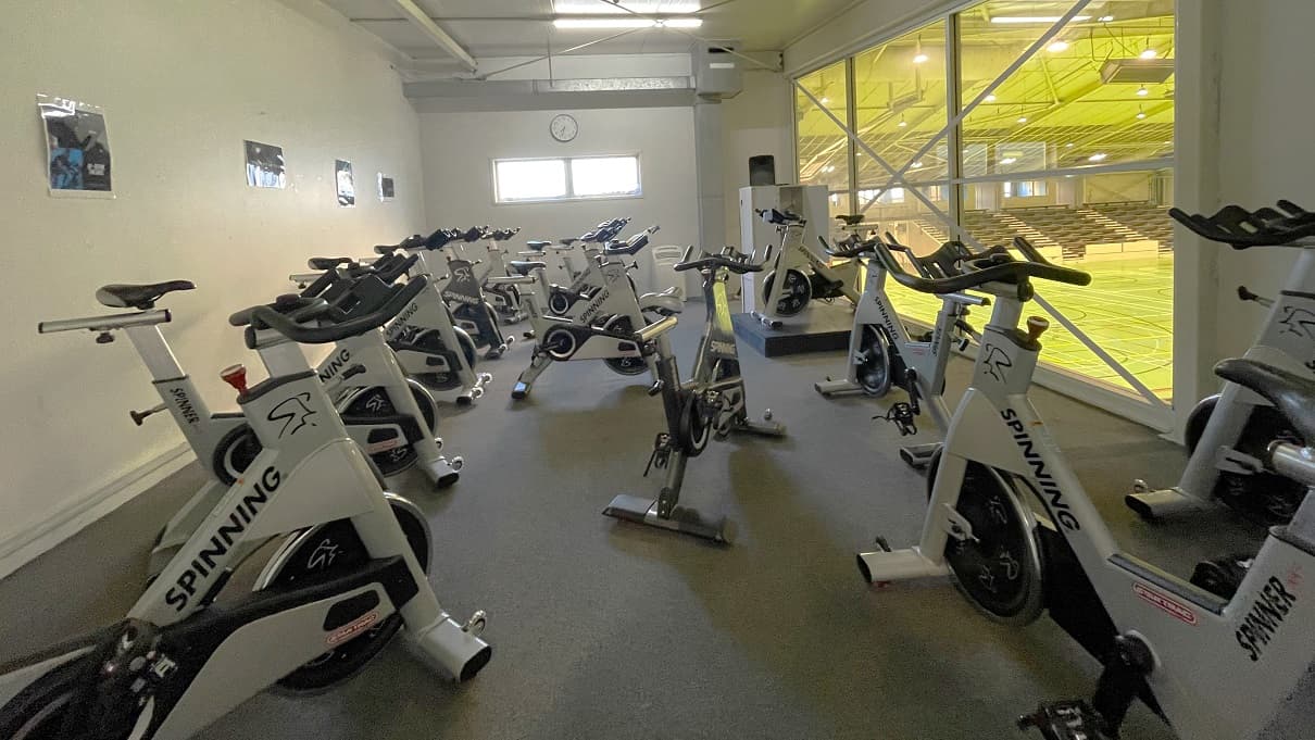 Studio 3 at the upper-level gymnasium in Trust House Recreation Centre. The studio has grey carpet tiles, a white painted wall, two small glass windows to the back, and a small clock on top of it. Several spin bikes are available at this studio. On the right wall is a floor-to-ceiling glass window that oversees the sports stadium