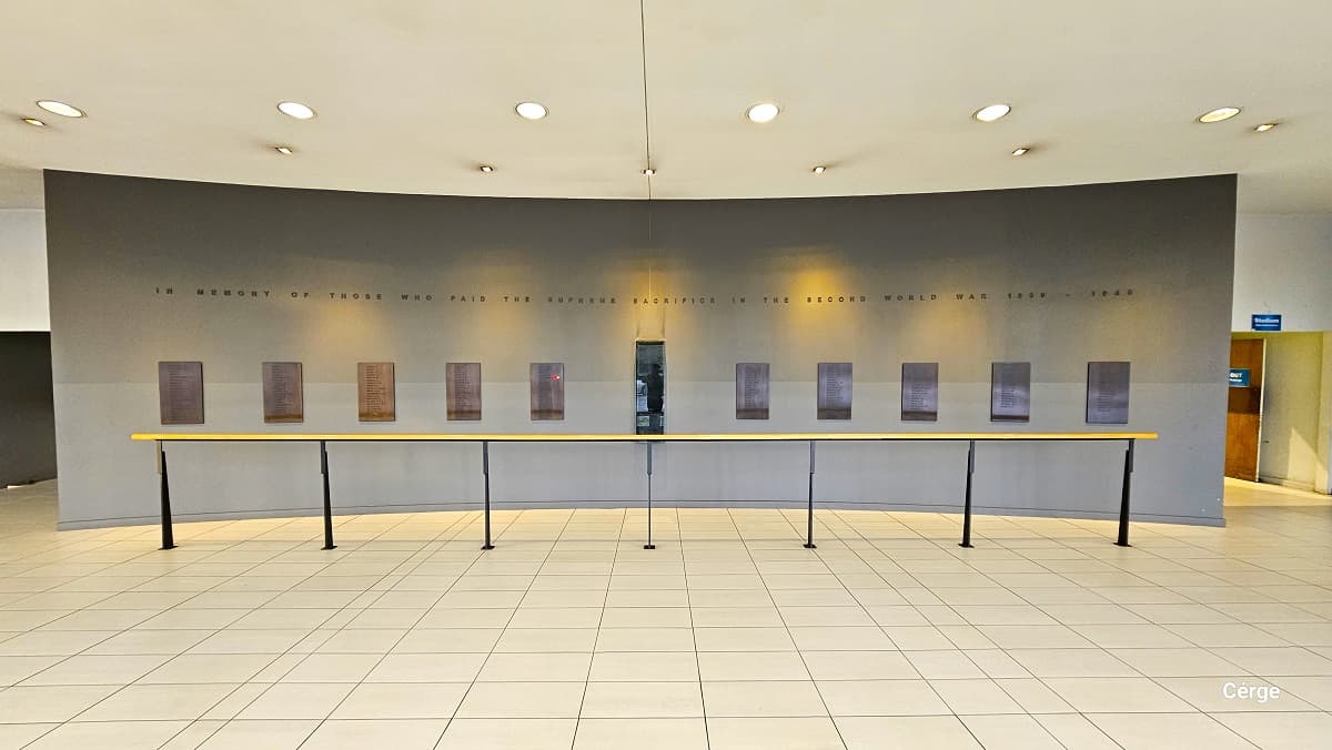 Hall of Honor at Trust House Recreation Center. On the right side is an outward opening, wooden entrance doors of the sports stadium, which is a large basketball court area. The hall has dirty white floor tiles and a white ceiling on the back of the grey curved wall with rectangular dark plaques. In front of it is a handrail to create a distance between guests and the hall. On the left grey wall is a large painting.
