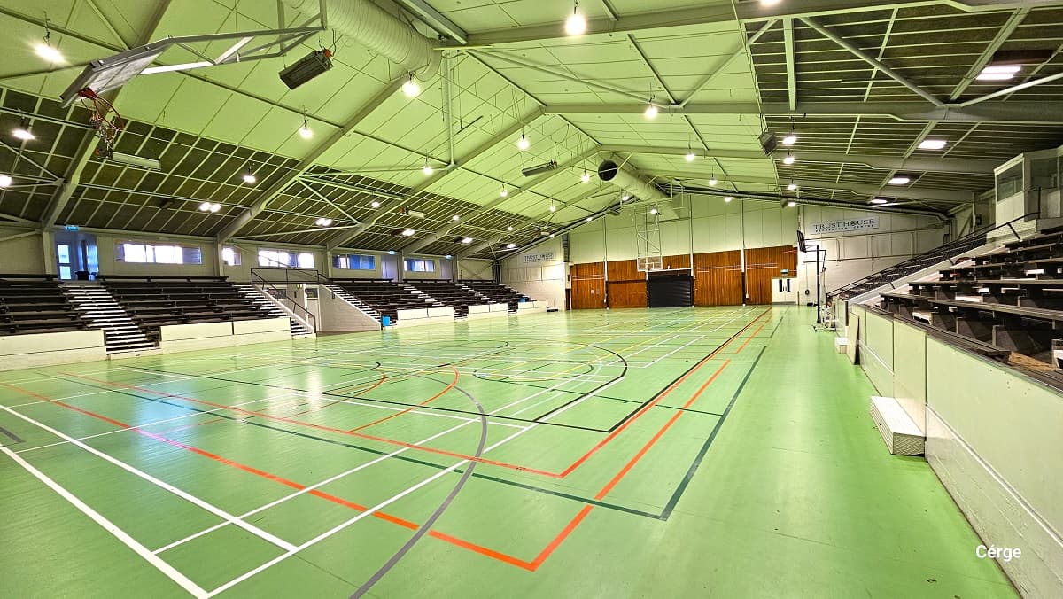 The basketball court has lime green timber floors with white, grey, black, orange, green, and yellow lines. The ceiling has a lime green ceiling with metal roofing frames and beams. The sports stadium has multiple black seating sections on each side of the court with a designated white walkway in between. In the middle of the seating is the dugout entrance with black handrails. On the back of the ring is a mahogany-painted wall.