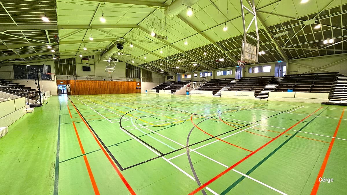 The basketball court has lime green timber floors with white, grey, black, orange, green, and yellow lines. The ceiling has a lime green ceiling with metal roofing frames and beams. The sports stadium has multiple black seating sections on each side of the court with a designated white walkway in between. The court is well-illuminated with artificial lighting.
