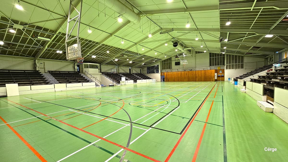 The basketball court has lime green timber floors with white, grey, black, orange, green, and yellow lines. The ceiling has a lime green ceiling with metal roofing frames and beams. The sports stadium has multiple black seating sections on each side of the court with a designated white walkway in between. On the back of the ring is a mahogany-painted wall and two doors on the left and right sides.