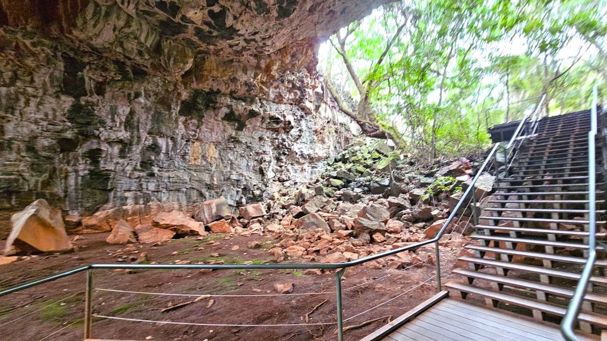 A rocky cave interior with a rough, textured wall on the left, covered in patches of moss and lichen. The ground is scattered with large, angular rocks and debris. On the right, a wooden staircase with metal railings ascends towards an opening surrounded by dense greenery and trees. The ceiling of the cave is uneven and jagged, with some overhanging sections. The floor is earthy and uneven, blending into the natural surroundings.