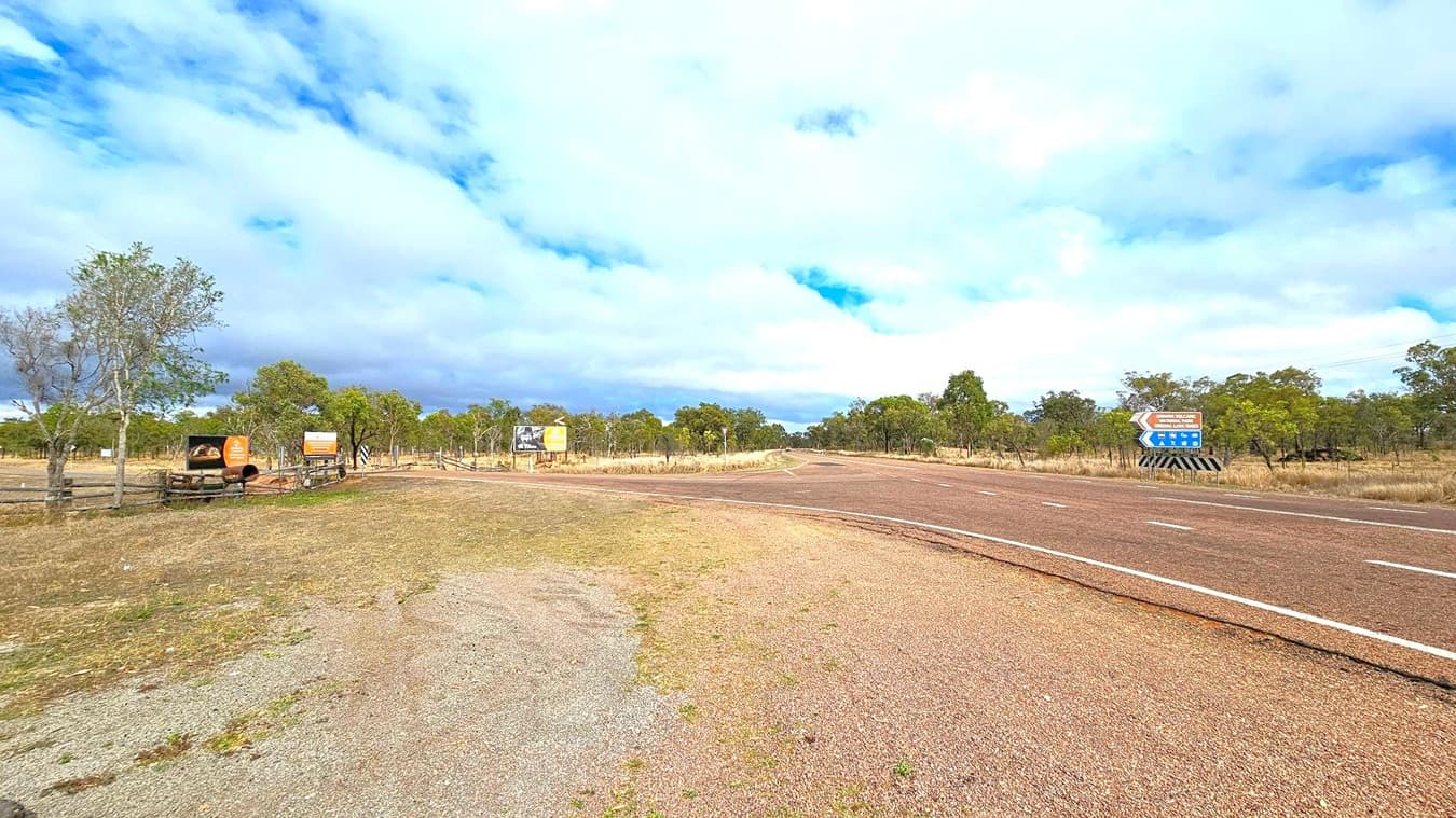 A rural road with a reddish-brown surface curves to the right. On the left, there are several signs, including one with an image of a cow and another with text. Sparse trees and grassland extend into the background. On the right, a blue directional sign with white text and arrows is visible. The sky is mostly cloudy with patches of blue. The ground is a mix of grass and gravel.
