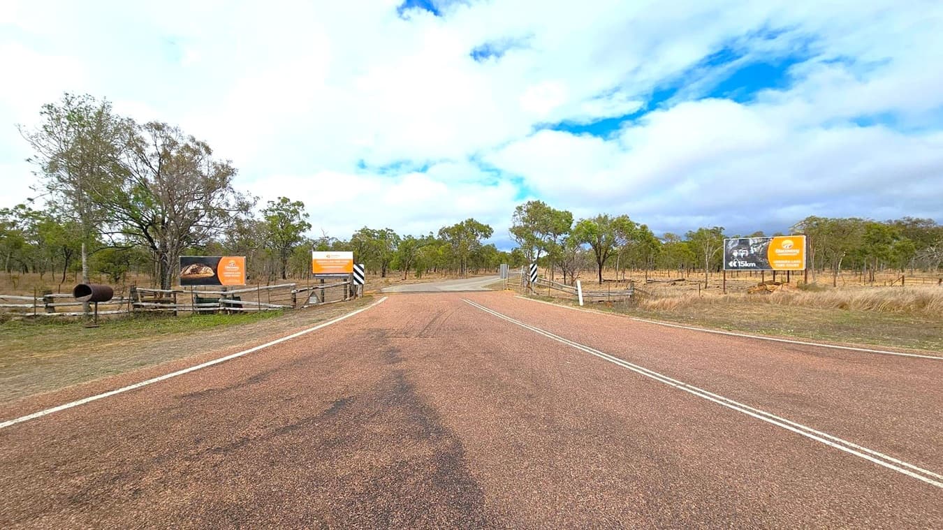 A rural road stretches into the distance, flanked by trees and grassland. On the left, a large tree stands near a black cylindrical object and two signs, one with an orange background. On the right, another orange sign is visible, along with a black and white striped post. The road is bordered by wooden fences on both sides, and the sky is overcast with patches of blue.