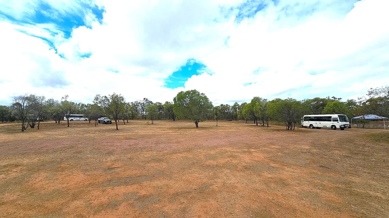 A wide, open field with sparse patches of grass and reddish-brown soil. In the background, there are several trees scattered across the landscape. On the left, a white bus is parked near some trees. In the centre, a few more trees are visible. On the right, another white bus is parked near a small structure with a canopy. The sky is mostly cloudy with some blue patches visible.