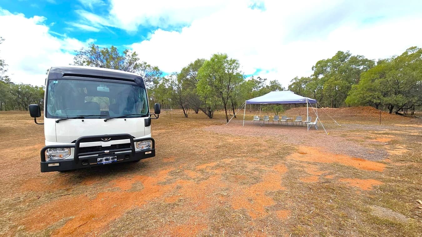 A white bus is parked on the left side on a patchy, reddish-brown dirt ground. On the right, there is a blue canopy tent with several chairs arranged underneath it. The background features scattered green trees and a mound of dirt. The sky is partly cloudy.