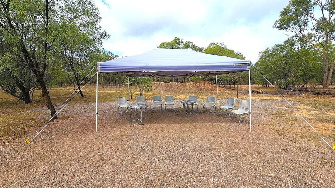 A large blue canopy tent is set up on a gravel surface. Underneath, there are several grey plastic chairs arranged in a semi-circle with a couple of small tables in the centre. The background features a natural setting with scattered trees and a mound of earth. The ground is a mix of reddish and grey gravel, and the tent is secured with ropes and stakes.