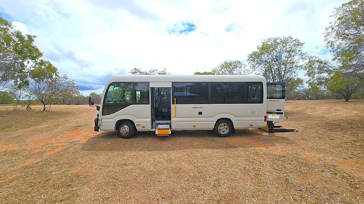 A white minibus is parked on a grassy area with patches of bare earth. The bus has its side door open, revealing a yellow step. At the rear, a platform is extended, possibly for wheelchair access. Surrounding the bus are scattered trees with sparse foliage. The sky is overcast, creating a muted backdrop.