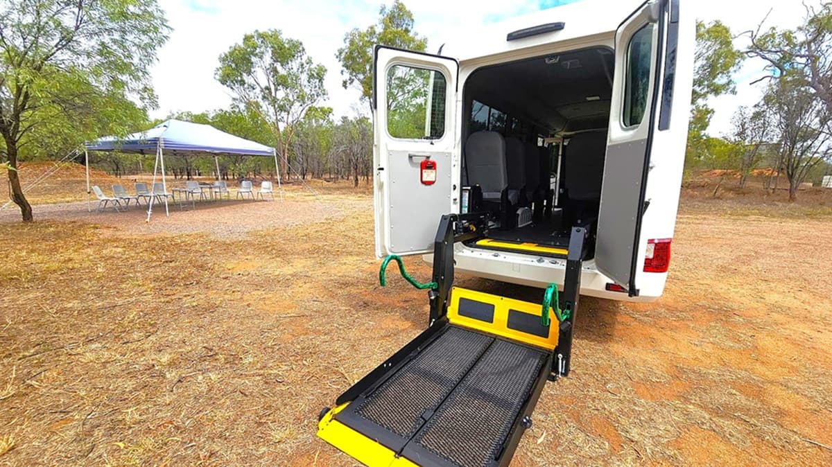 A white van with its rear doors open, revealing a yellow and black wheelchair lift extended onto the ground. The van is parked on a dry, grassy area. To the left, a blue canopy tent is set up with several chairs arranged around a table underneath. Trees with sparse foliage are scattered in the background, and the ground is covered with dry grass and patches of dirt.