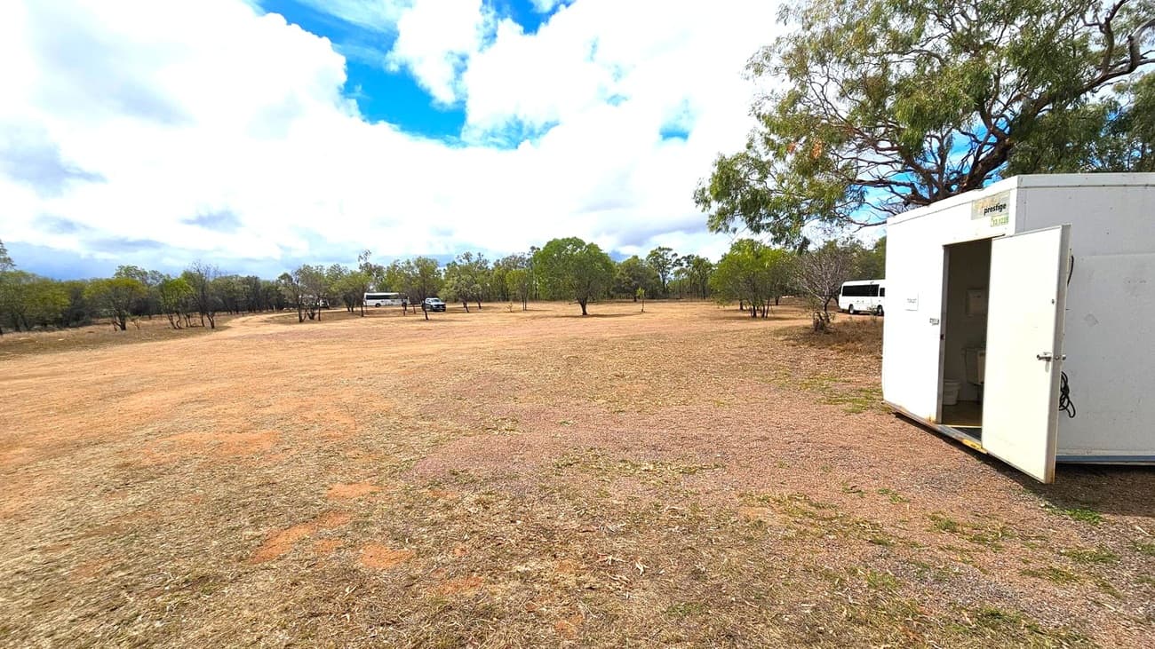 A wide, open field with sparse grass and patches of bare earth. On the right, a white portable building with an open door. In the background, several trees are scattered across the landscape. Two white vehicles, including a bus, are parked near the trees. The sky is filled with large, fluffy clouds.